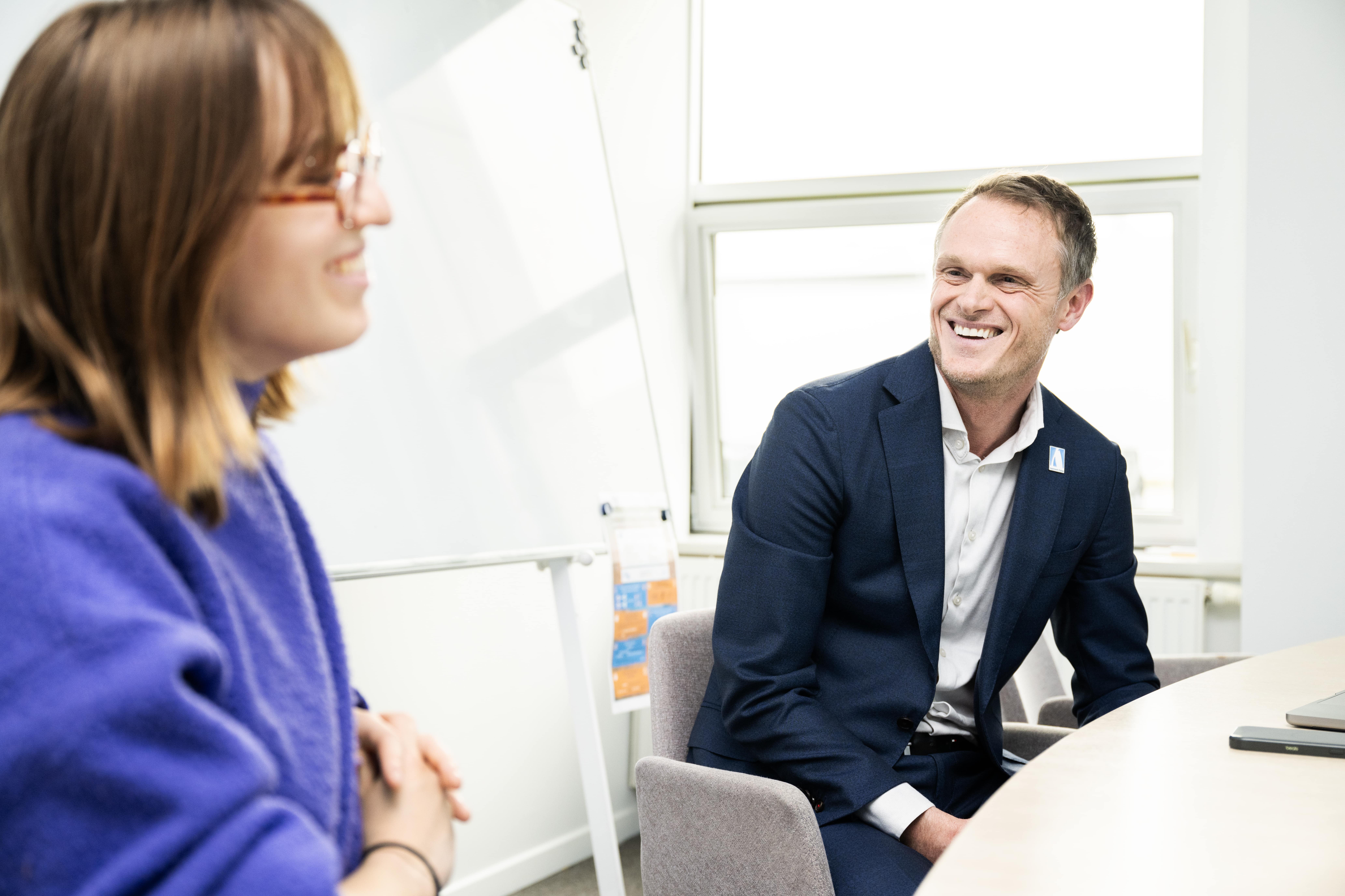 Man in a suit smiling during a meeting with a woman in a blue sweater, sitting at a conference table