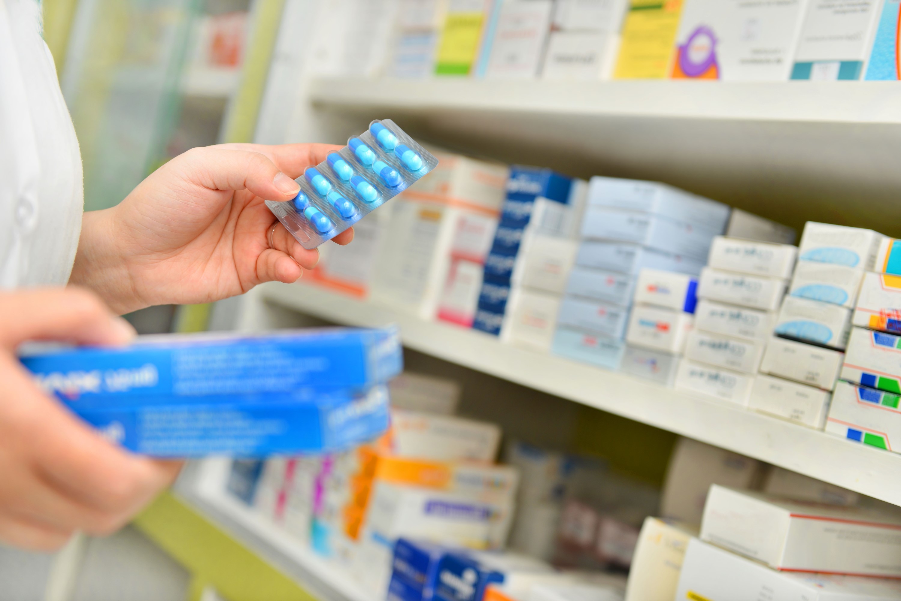 Hands selecting blue blister pack medicine from pharmacy shelf with boxes of medication in the background