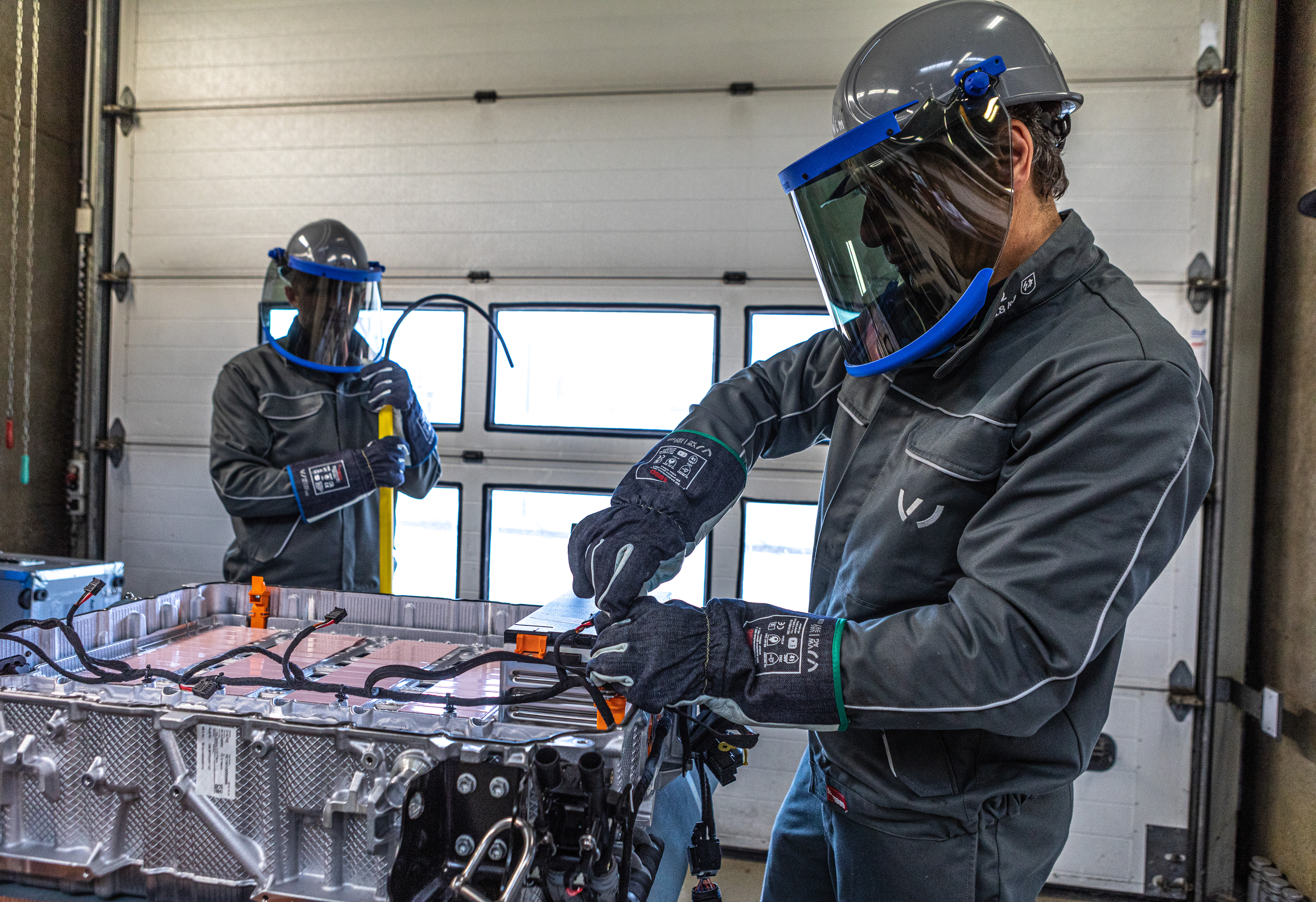 Two technicians in protective gear work on a large battery pack inside a garage, with one person using tools and the other holding a yellow handle.