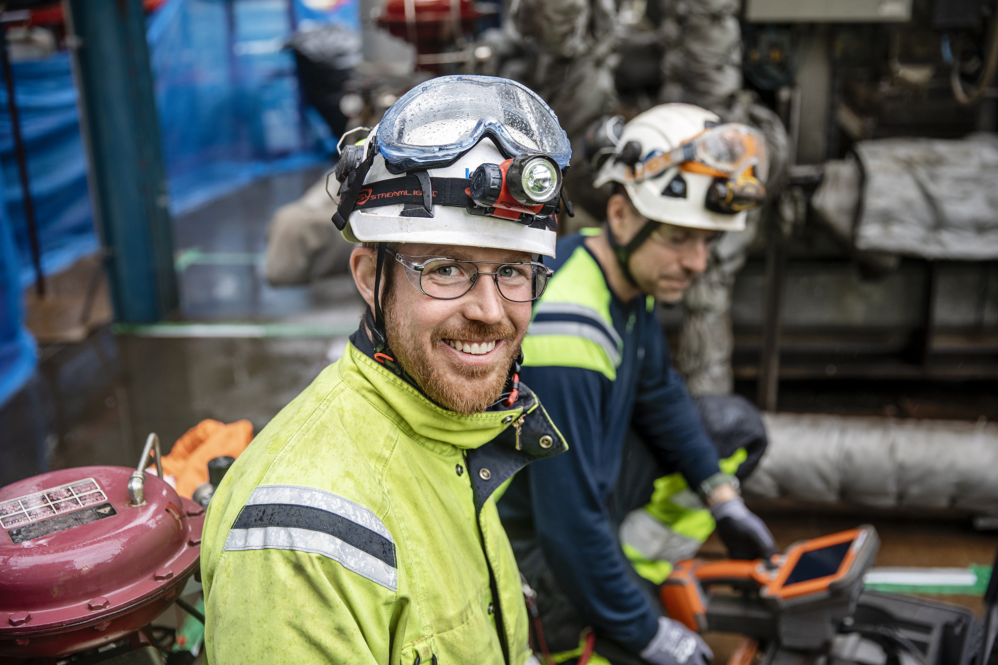 Smiling Kiwa auditor wearing safety gear, including a hard hat and safety glasses, in an industrial setting with machinery in the background