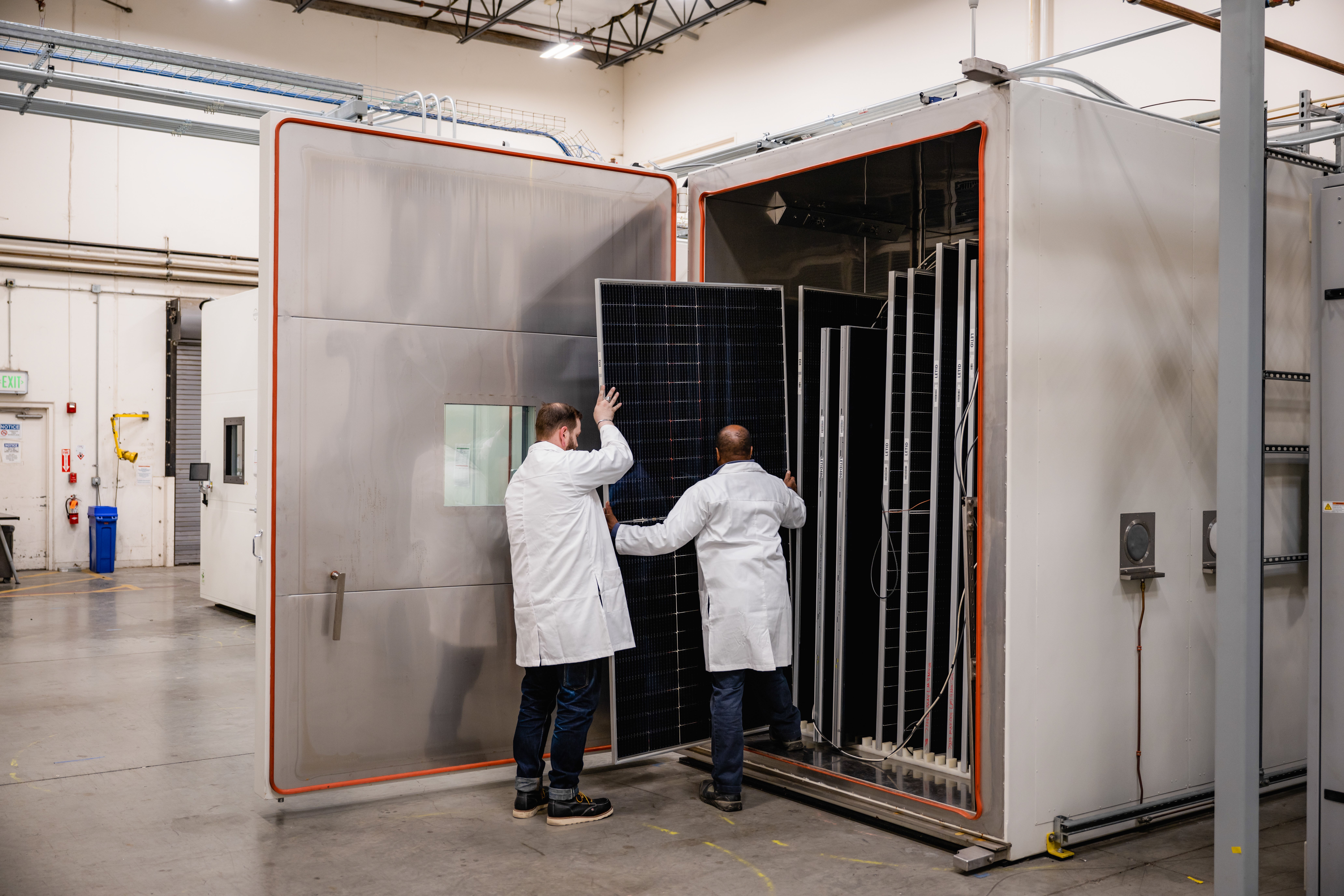Two Kiwa PVEL laboratory employees in white lab coats handling a large solar panel inside an industrial testing chamber.