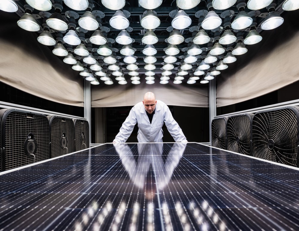 Kiwa PVEl laboratory employee in a white lab coat standing behind a large solar panel under an array of bright industrial lights in a testing chamber.